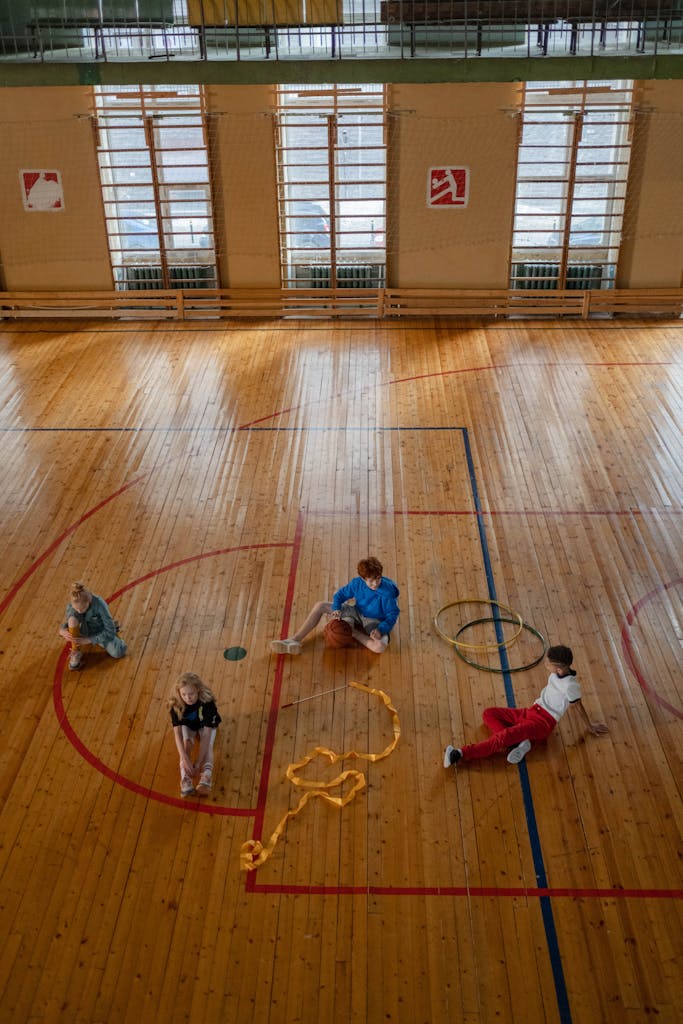 Kids are engaging in activities on a wooden sports hall floor with hula hoops and ropes.